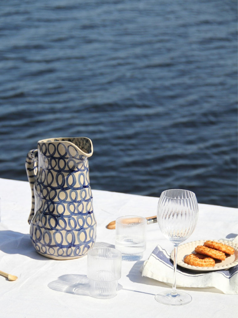 A patterned ceramic pitcher with blue and beige geometric circles sits on a white tablecloth outdoors with the ocean water in the background, alongside drinking glasses and a plate of food.