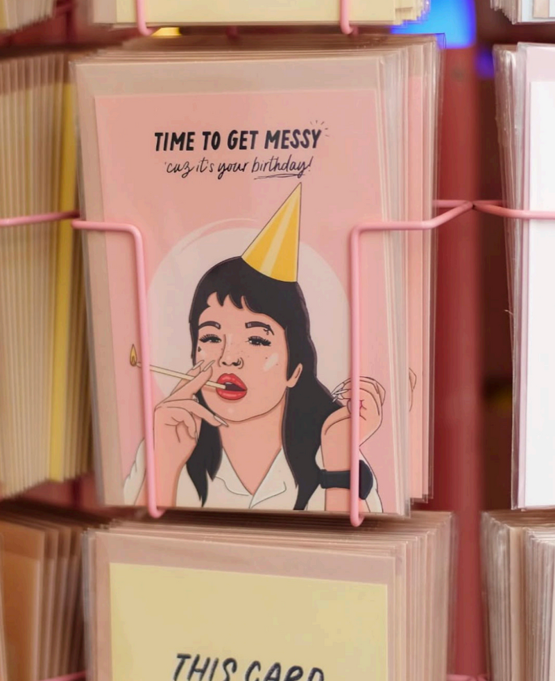 A pink greeting card displayed in a pink wire rack, featuring an illustration of a woman looking like Lola Young with dark hair wearing a party hat and holding a cigarette, with the text 'TIME TO GET MESSY catch it's your birthday!'.