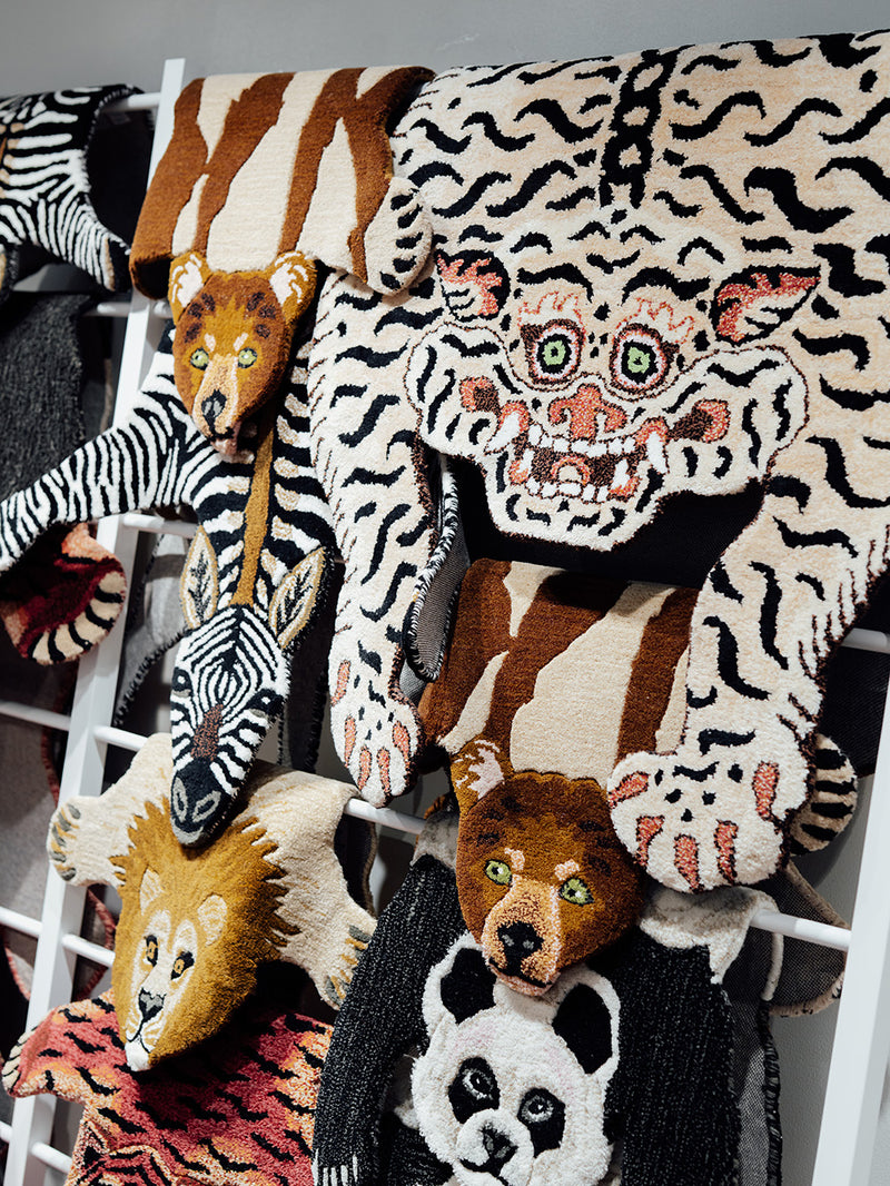 A close-up of several animal-themed rugs hanging on a white rack. The designs include a tiger face, zebra, lion, bear, panda, and other animal patterns in black, white, brown, beige, and some pink accents.
