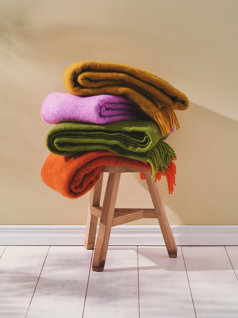 Stack of colorful folded blankets on a wooden stool against a beige wall.