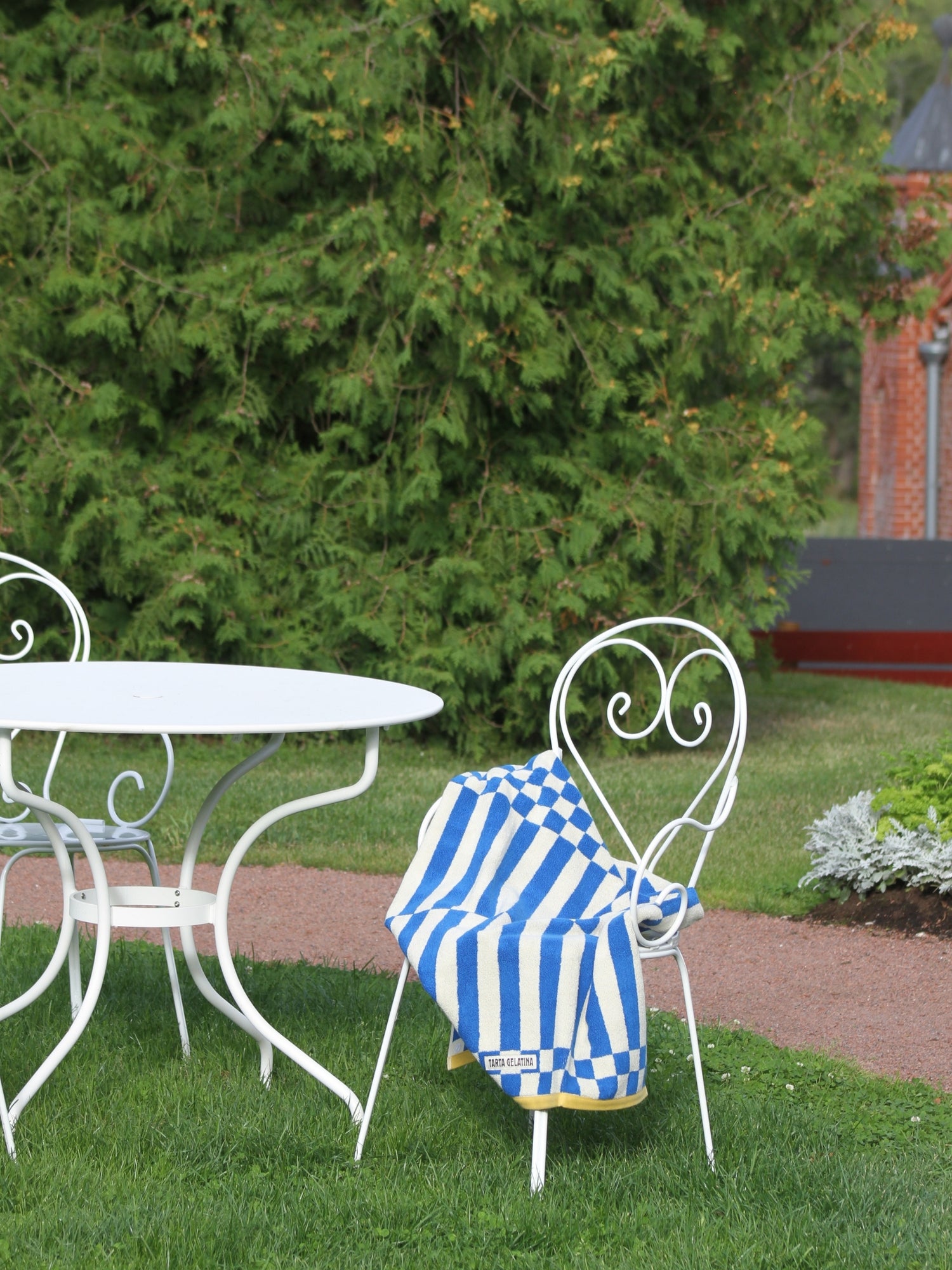 A blue and white striped towel with a gold border is draped over a white wrought-iron garden chair in an outdoor backyard setting with a white table, green lawn, and large green shrubs.