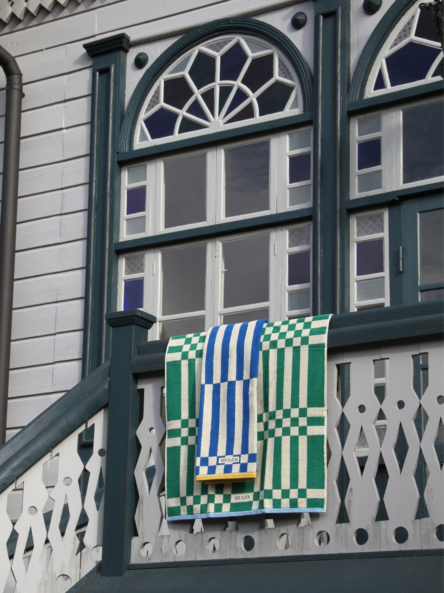 A green and white checkered towel with blue and white striped patches hanging from a dark green wooden balcony railing of a house with white siding and decorative windows.