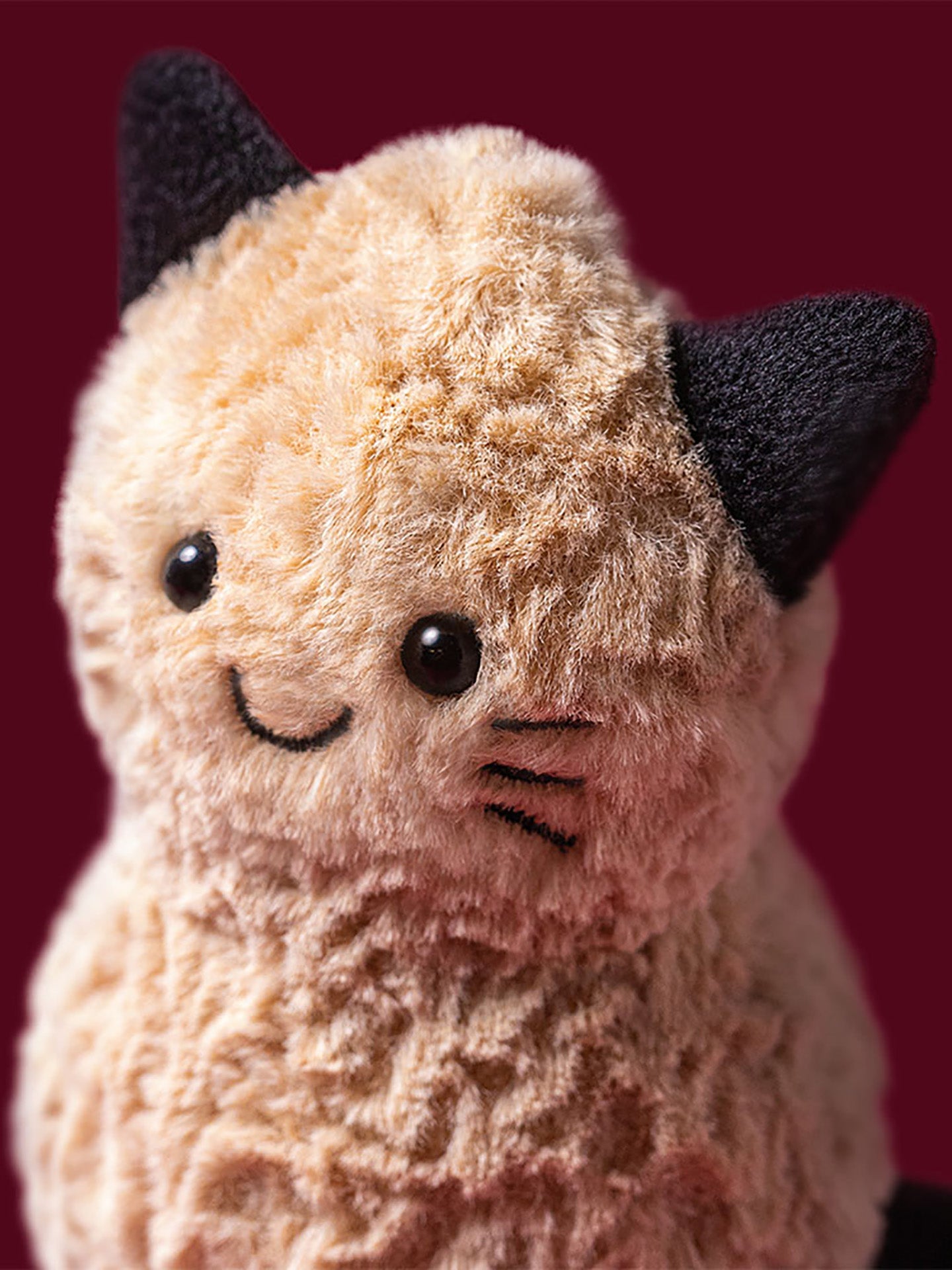 A close-up of a plush peanut soft toy with cream-colored fuzzy fur, dressed in black cat halloween ears, and a stitched smiling face with cat whiskers against a dark burgundy background.