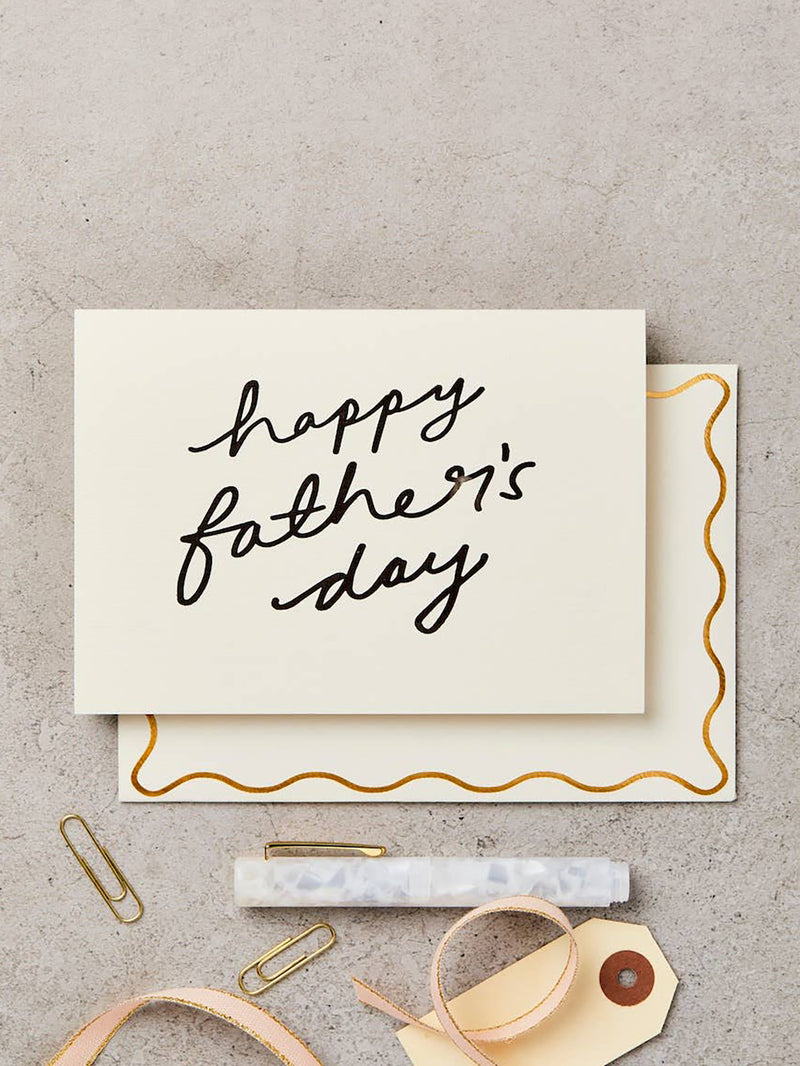 A Father's Day greeting card with 'happy father's day' written in black script, layered over a matching cream envelope with a gold wavy border. The card and envelope are arranged on a textured gray surface with gift accessories including paper clips, a marble pen, and a ribbon tag.