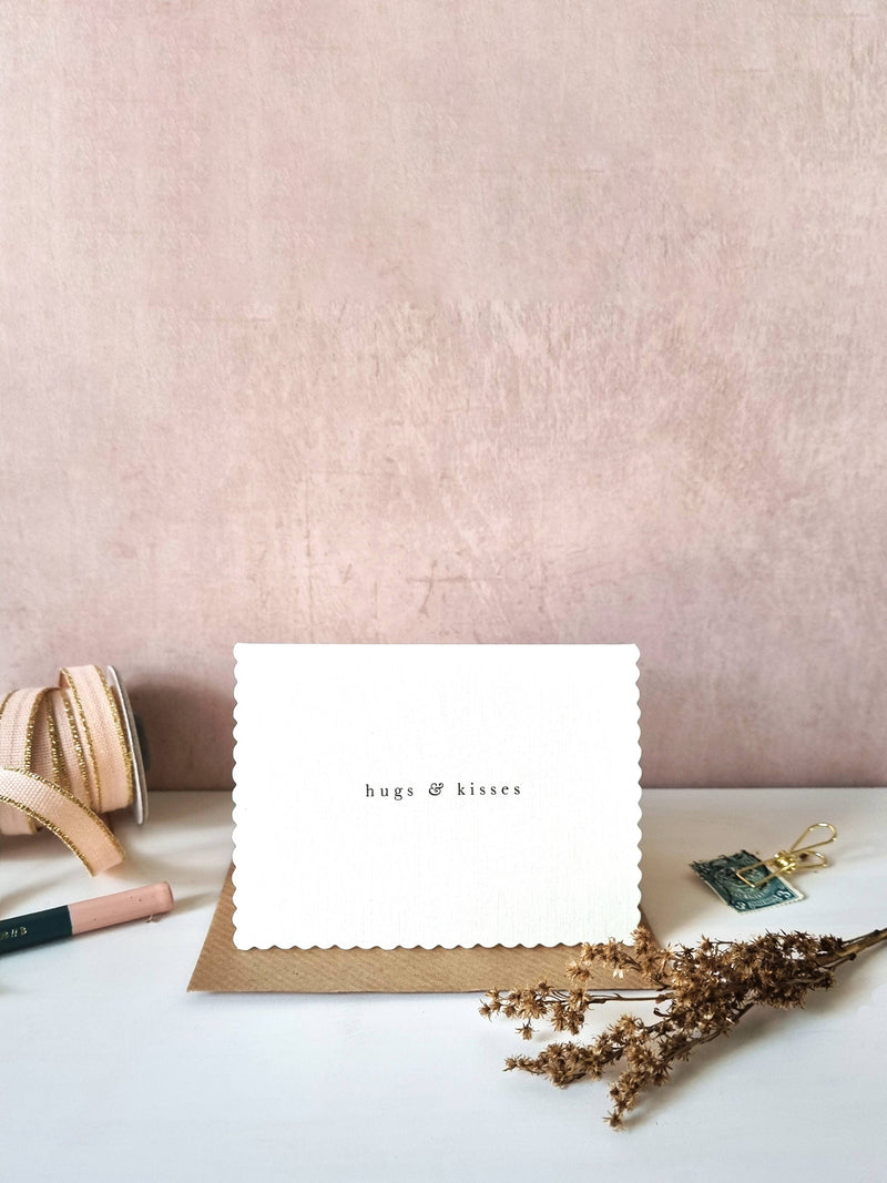 A white scalloped-edge greeting card with the words 'hugs & kisses' printed in small black text, resting on a brown kraft envelope on a white tabletop. The scene is styled with dried flowers, a spool of ribbon, and a small decorative tag against a textured dusty pink wall.
