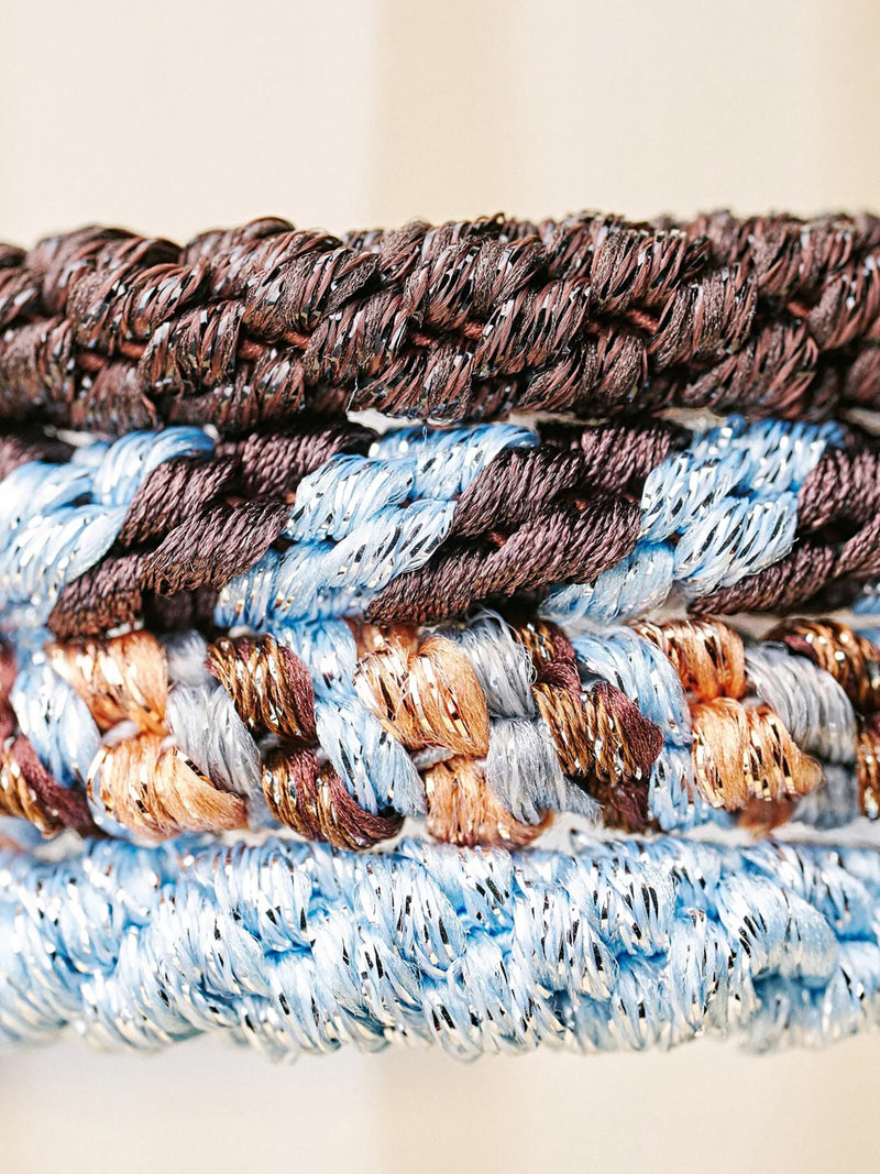 Close-up of 4 stacked braided hair ties or ponytail holders all different but matching colours, including two solids, a dark brown and a light blue, with 2 mixed colours, one with peach, light blue, light grey and brown combination and other one with only light blue and brown mixture, all spun with little metallic silver thread running with the braiding.
