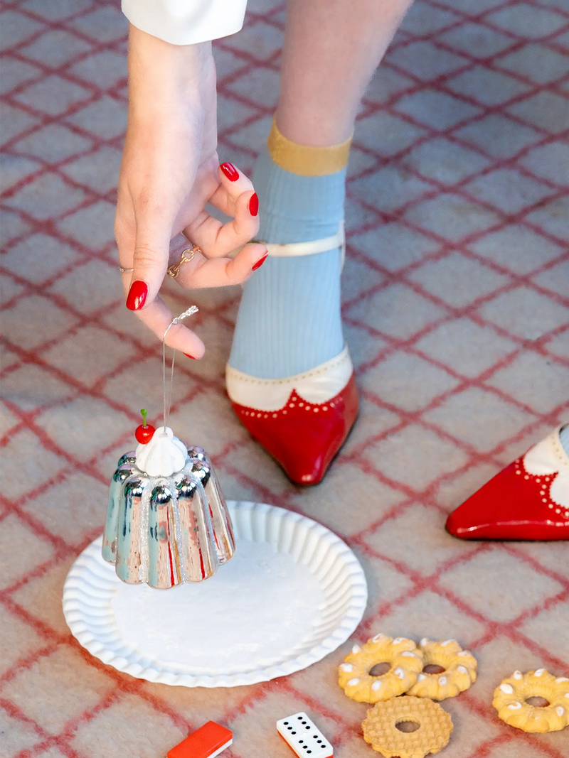 A person's hand with red nail polish holds a miniature glass pudding ornament above a white paper plate. The person is wearing light blue and red patterned Mary Jane socks, standing on a red-and-white checkered carpet.