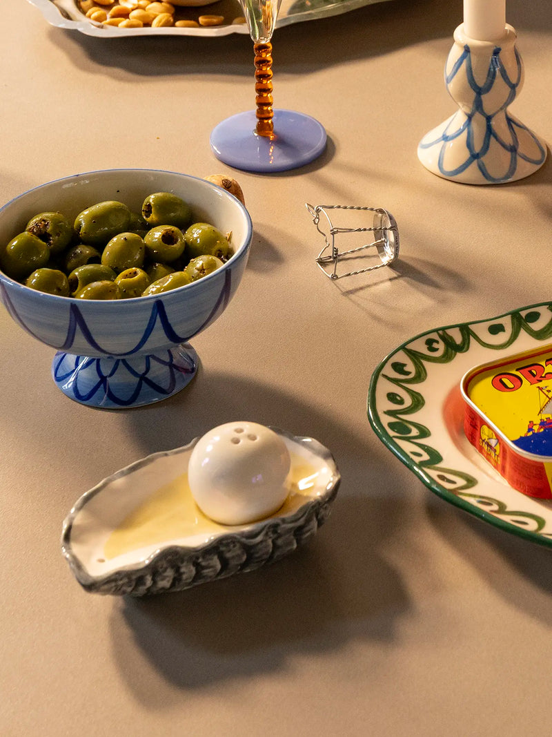 A styled tabletop arrangement featuring a blue and white patterned ceramic bowl of green olives, a matching blue and white pedestal plate, a gray clam-shell style dish holding a white pearl, and other decorative tableware on a beige surface.