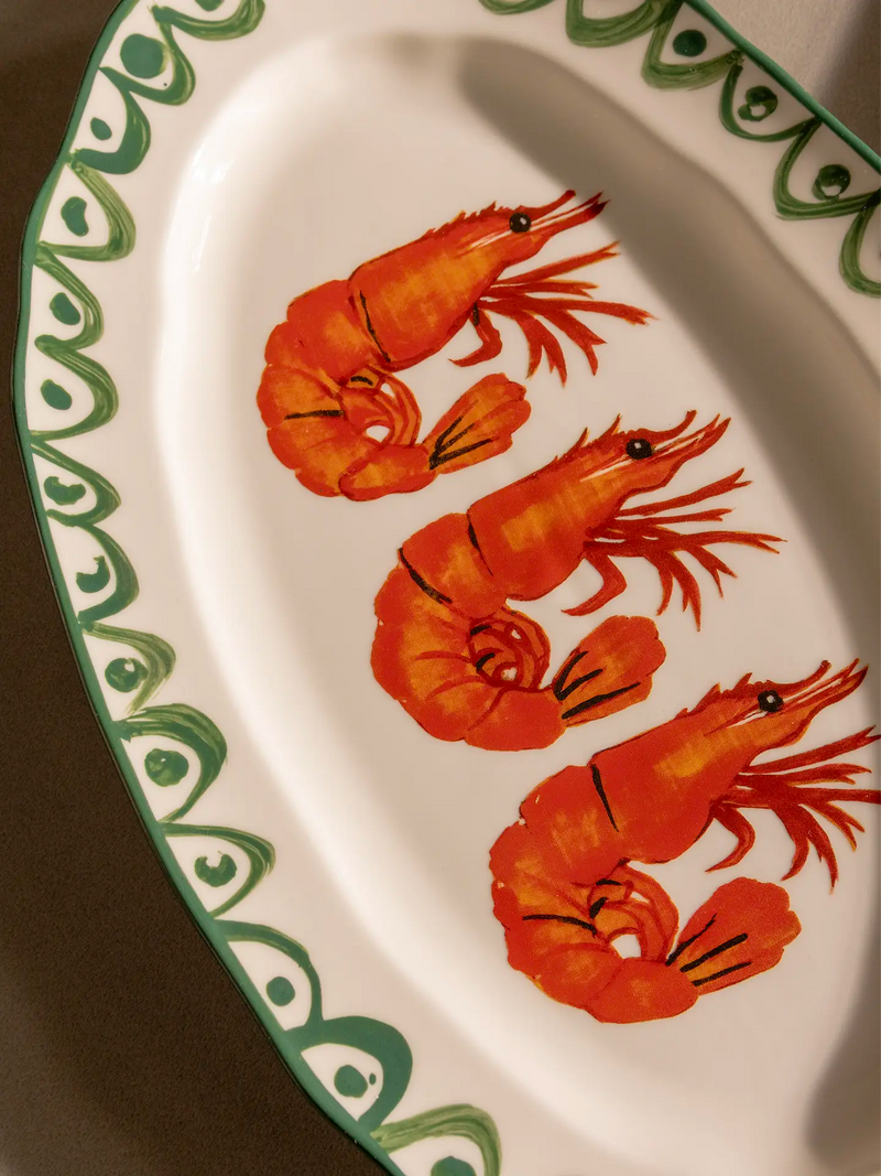 An oval white ceramic serving platter with a green, hand-painted scalloped border decoration and three painted orange-red prawns, placed on a dark brown surface and cropped for a close-up.