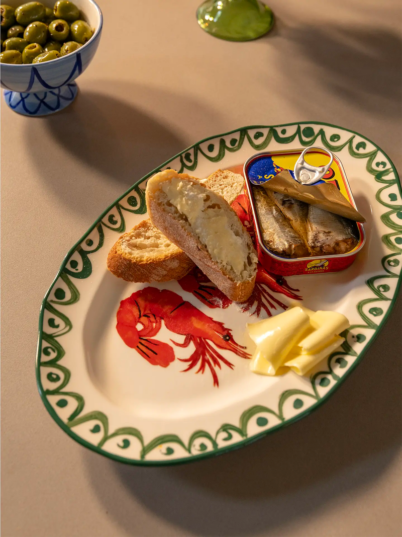 An oval ceramic serving platter with a green patterned border and a red shrimp illustration, styled with sliced bread, butter, a can of sardines, and a bowl of olives on a beige tabletop.