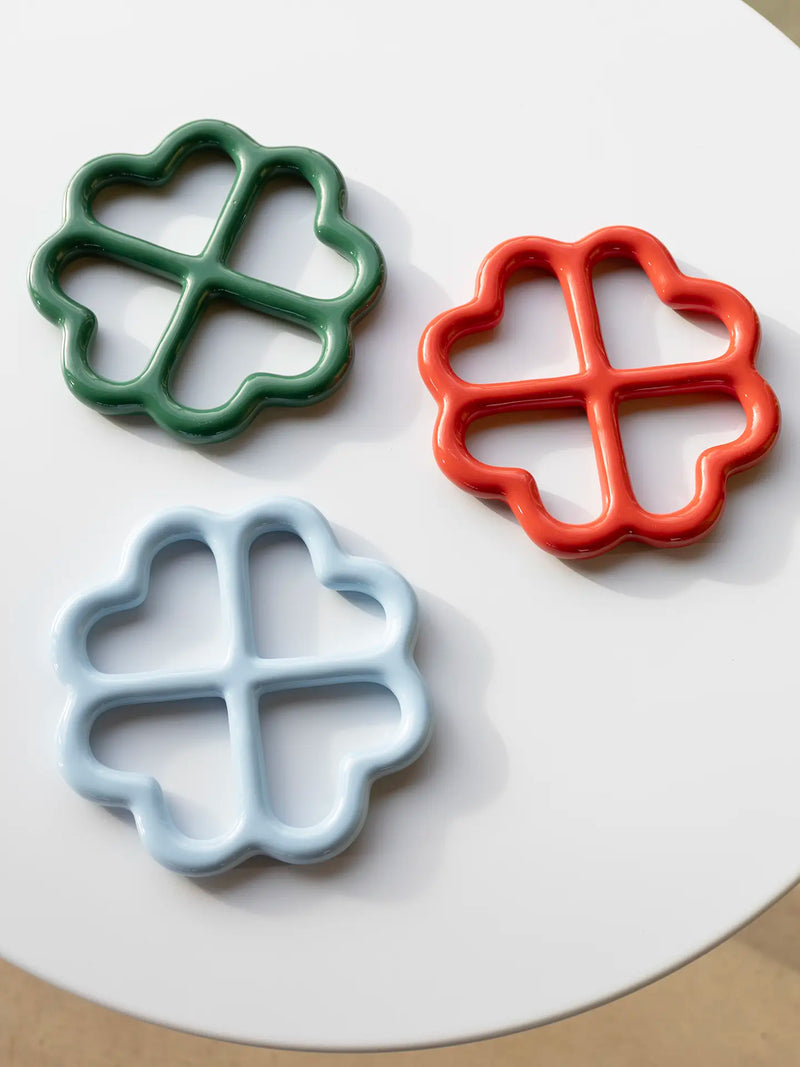 Three flower-shaped ceramic coasters or trviets in green, red, and light blue, arranged on a white round table.