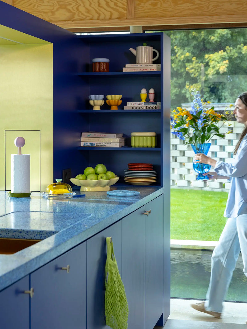 A woman in a light blue outfit walks into a light-filled colourful modern kitchen carrying a blue glass vase filled with yellow and blue flowers. The kitchen features deep navy blue cabinets, a blue speckled countertop, and open shelves displaying colorful dishes, such as butter yellow wavy bowl filled with apples and a pastel blue trivet on the countertop and stylish cookbooks on the shelf.