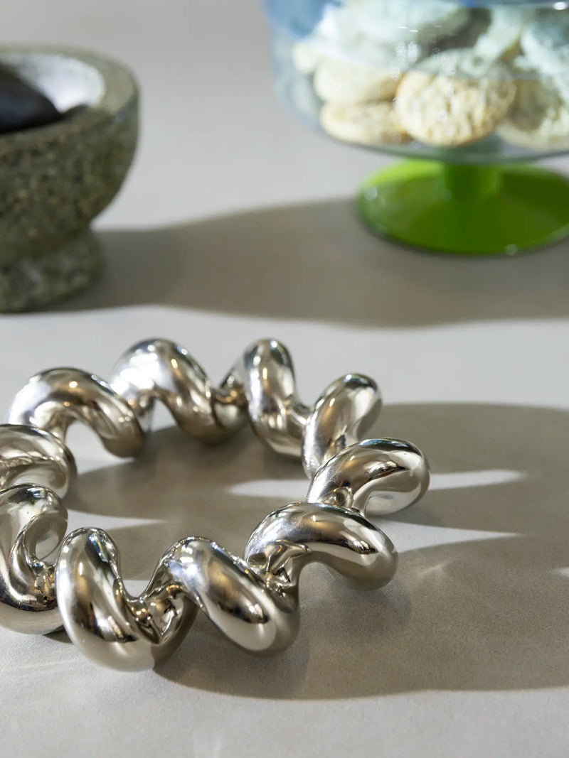 A chunky metallic silver trivet with a bold, wavy, linked design, displayed on a light beige surface with other kitchen items on soft-focus background.