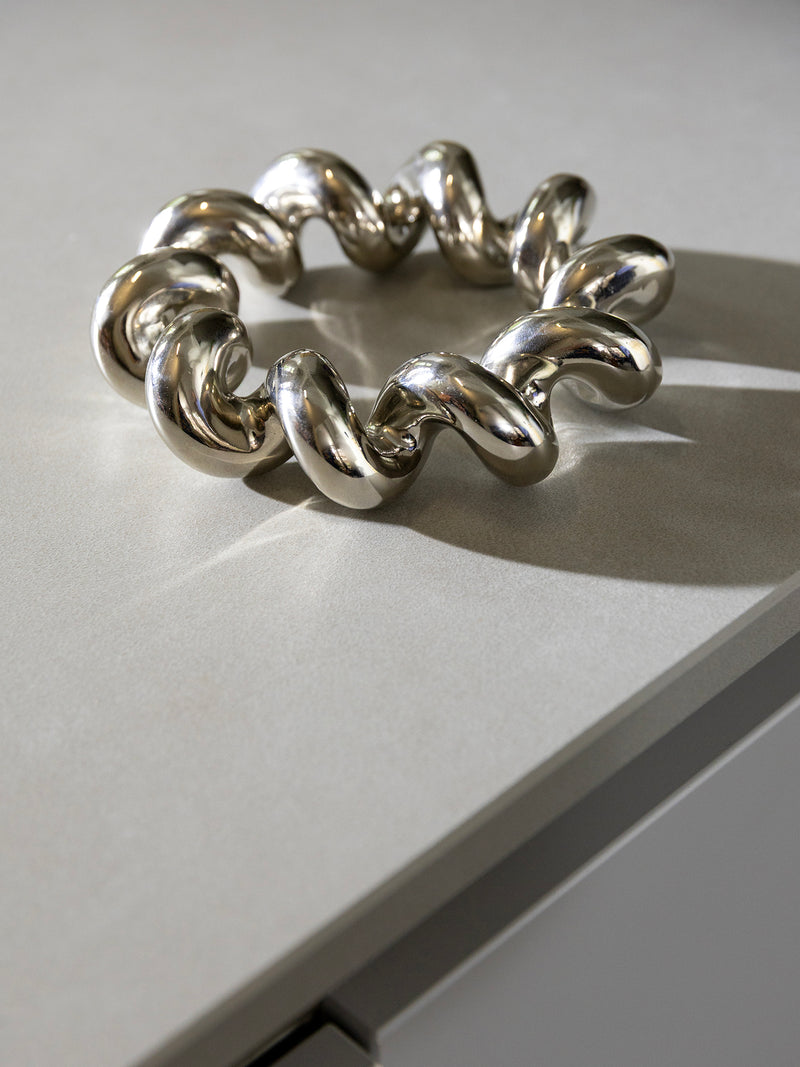 A chunky, polished silver-tone metal-looking trivet with a wavy, spiral design, displayed on a light gray surface.