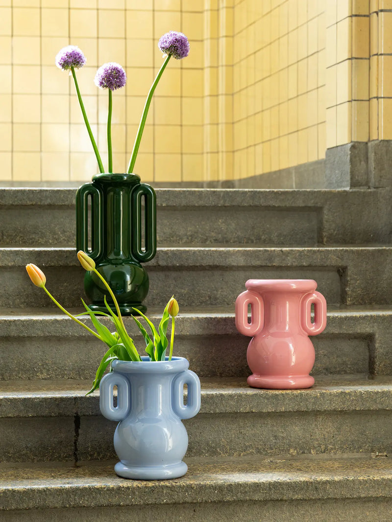 Three ceramic amfora or urn-style flower vases in dark green, cool light blue, and sweet pink are arranged on stone steps against a yellow tiled wall. The light blue vase holds tulips, and the dark green vase holds allium-like purple flowers.