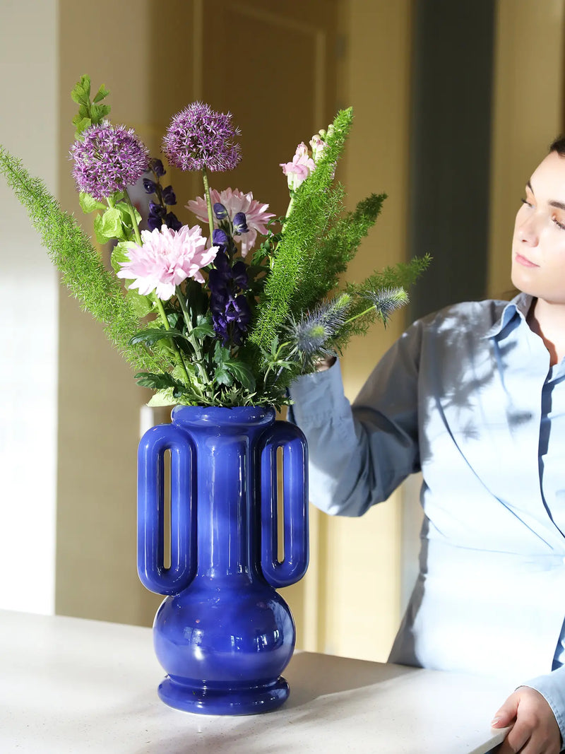 A person in a light blue shirt adjusts a large cobalt blue ceramic vase filled with pink and purple flowers and green foliage on a tabletop in an indoor setting.