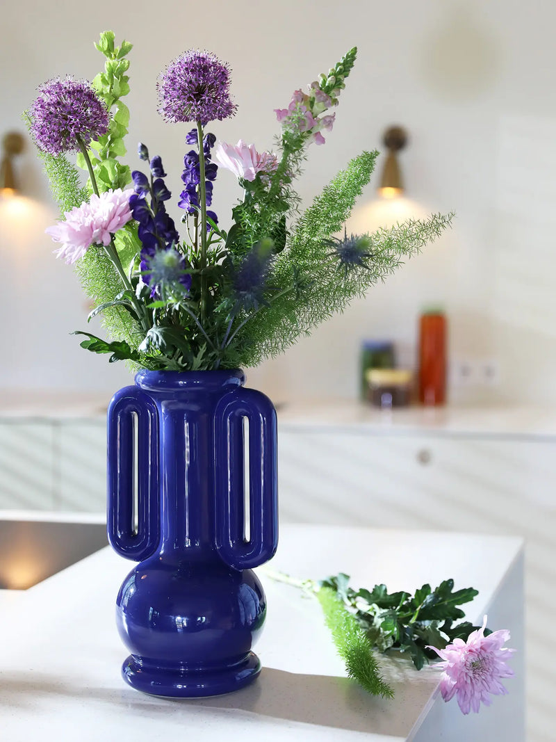 A glossy cobalt blue ceramic vase with two vertical handles, filled with a colorful floral arrangement of purple, pink, and green flowers, set on a white countertop in a softly lit kitchen.