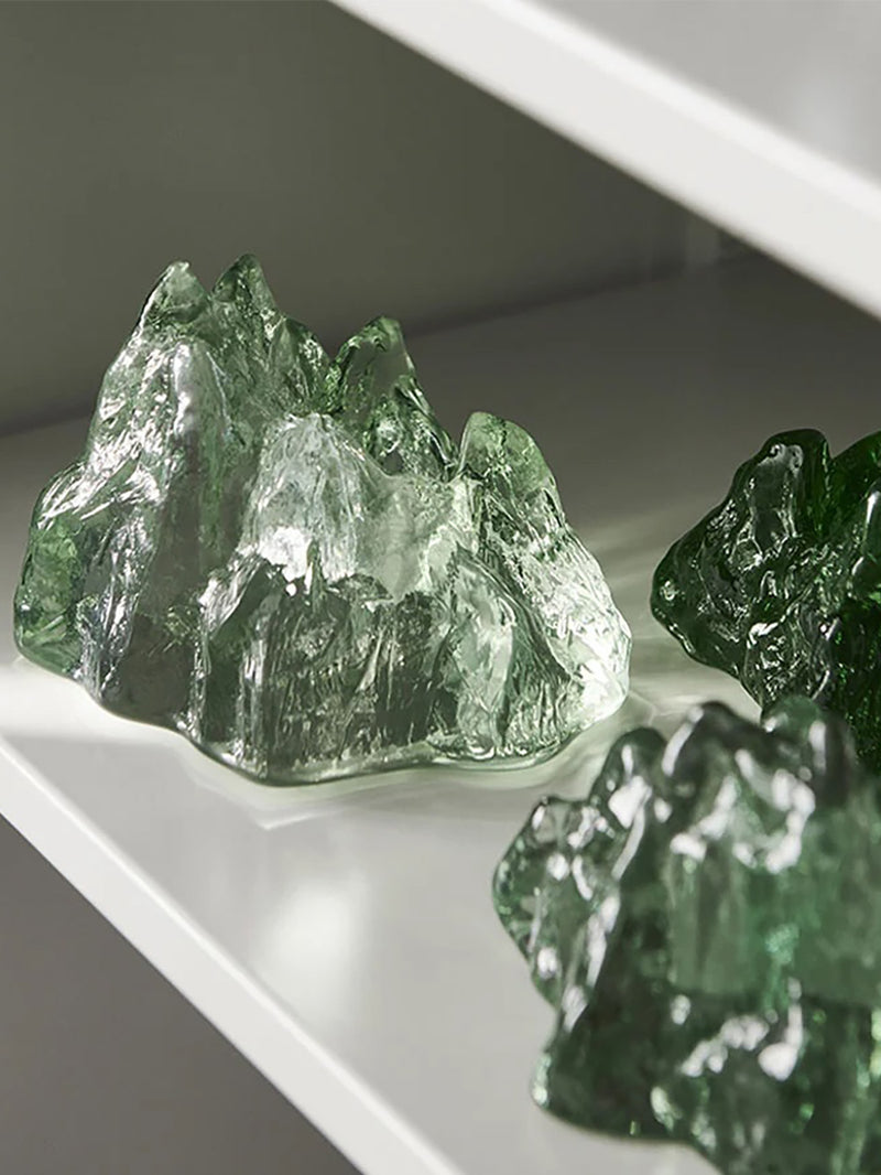 Three sculptural glass mountain-like tealight holders in different green tones arranged on a white shelf.