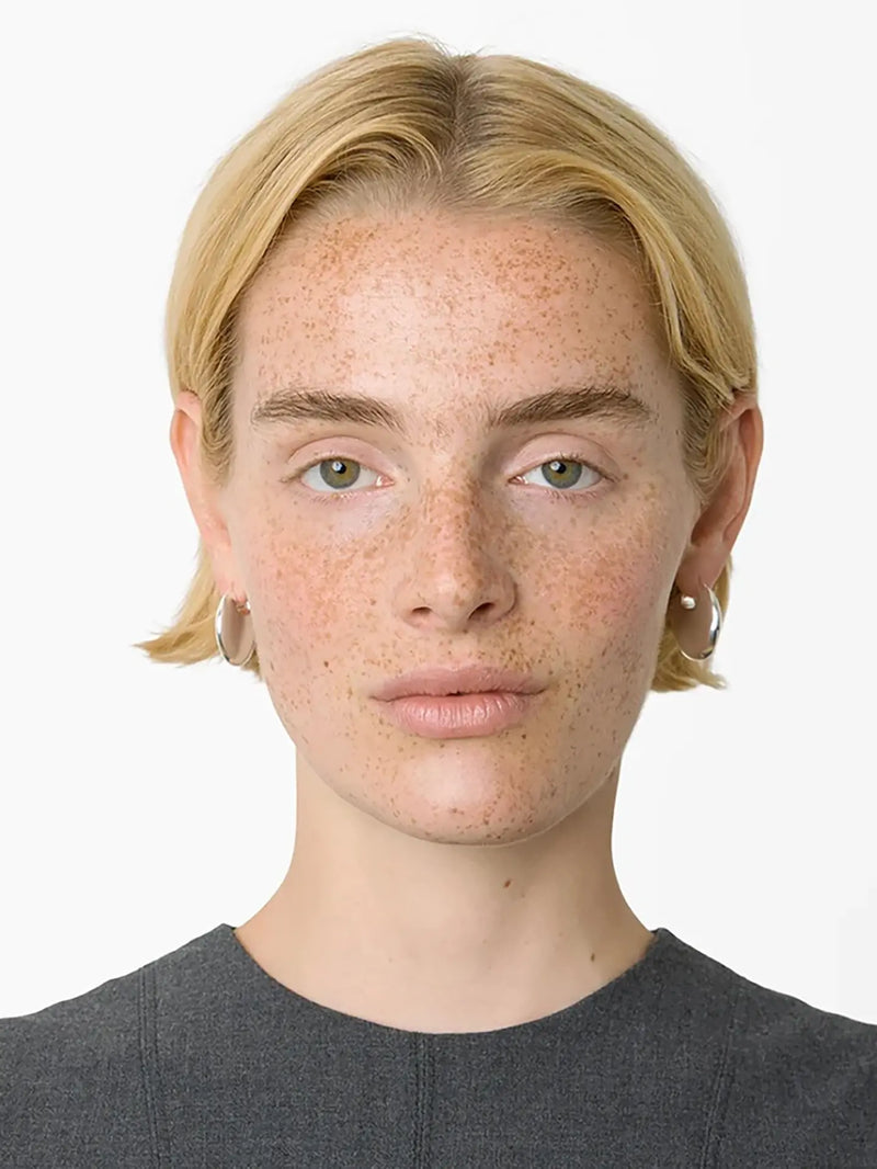 A close-up portrait of a person with short blonde hair and freckles, wearing small silver hoop earrings and a dark gray top against a plain white background.