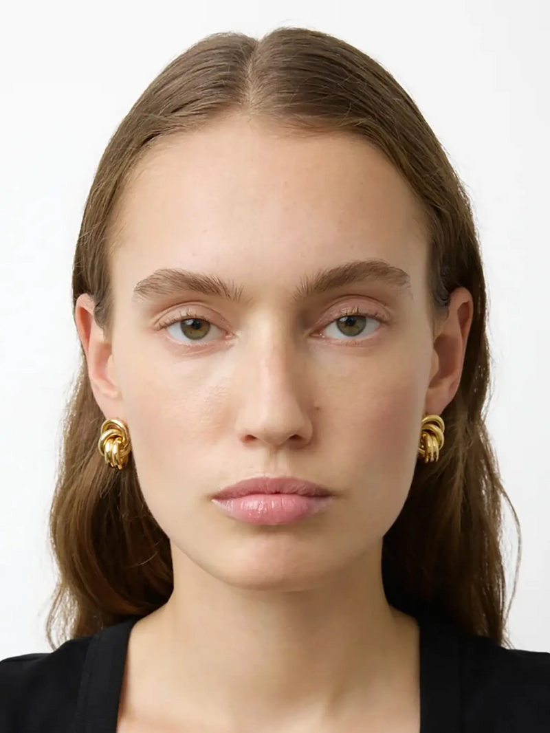 A close-up portrait of a woman with brown hair wearing gold knotted hoop earrings against a plain white background.