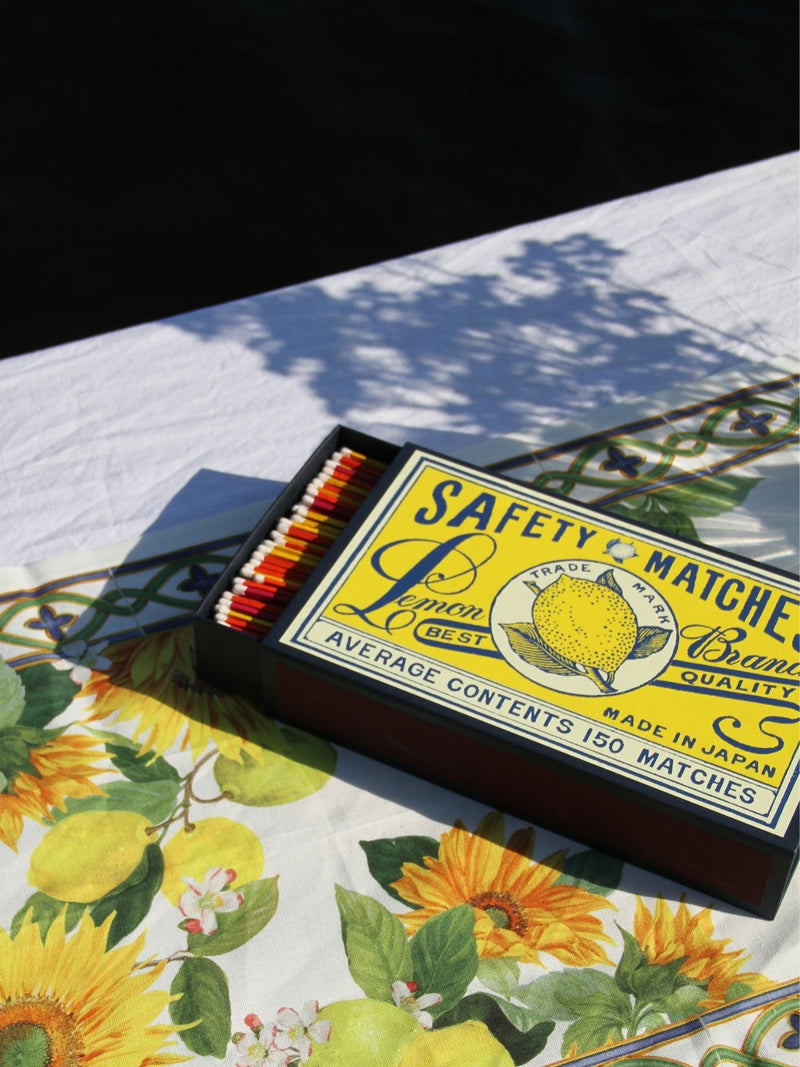 A vintage-style matchbox labeled 'Safety Matches Lemon Balm Quality' lies open on a floral tablecloth with sunflowers and green leaves. The matchbox is dark brown with a yellow label and red-and-white matches inside.