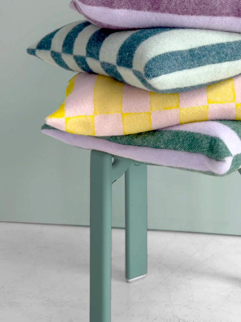 A stack of four patterned throw pillows in pastel colors sits on a mint green table or chair stanging on marble floor and photographed against a pale green wall.