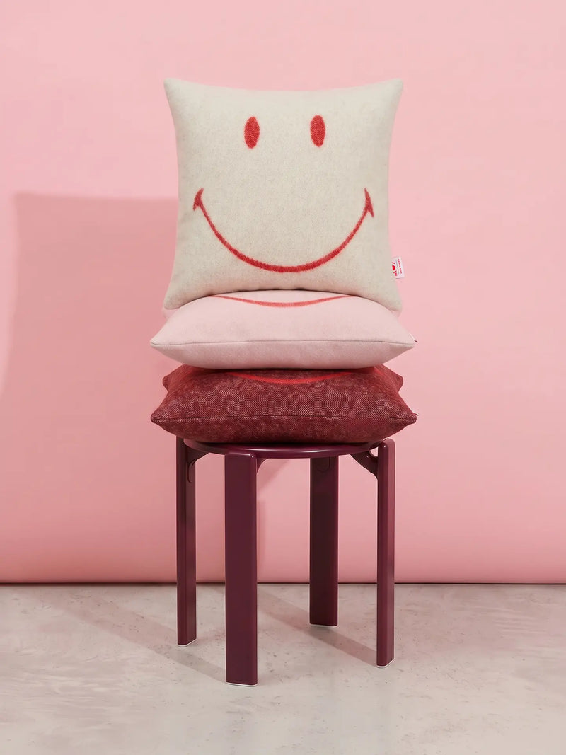 Three stacked decorative throw pillows in cream, pale pink, and dark red, each with a simple red smiley face design, displayed on a dark red stool against a solid pink wall.