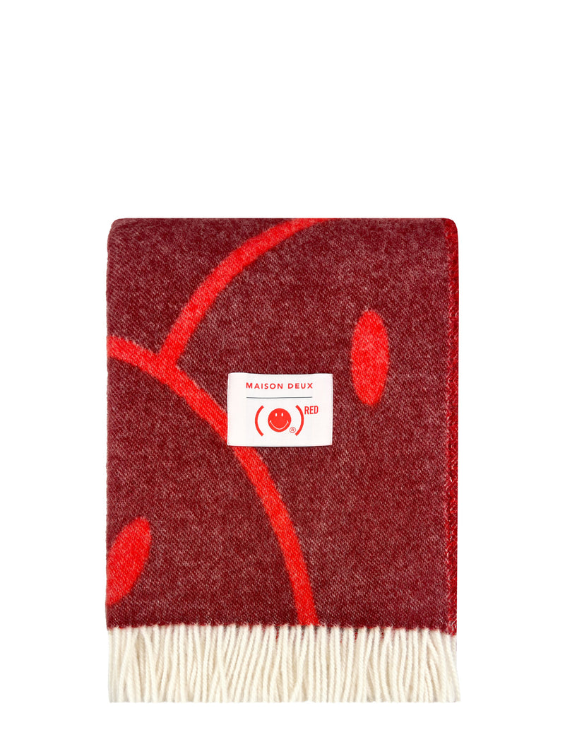 A Maison deux throw blanket in a deep burgundy red, featuring a bright red abstract leaf pattern and a white woven label in the center, finished with cream-colored fringe at the bottom.