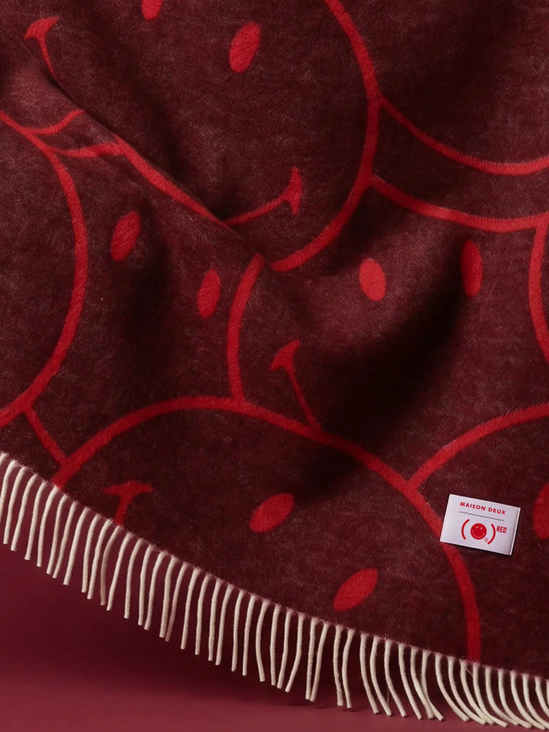 A close-up of a dark burgundy throw blanket featuring a red smiley face pattern and cream-colored fringe along the edge, with a small white 'Maison Robert' brand tag visible.