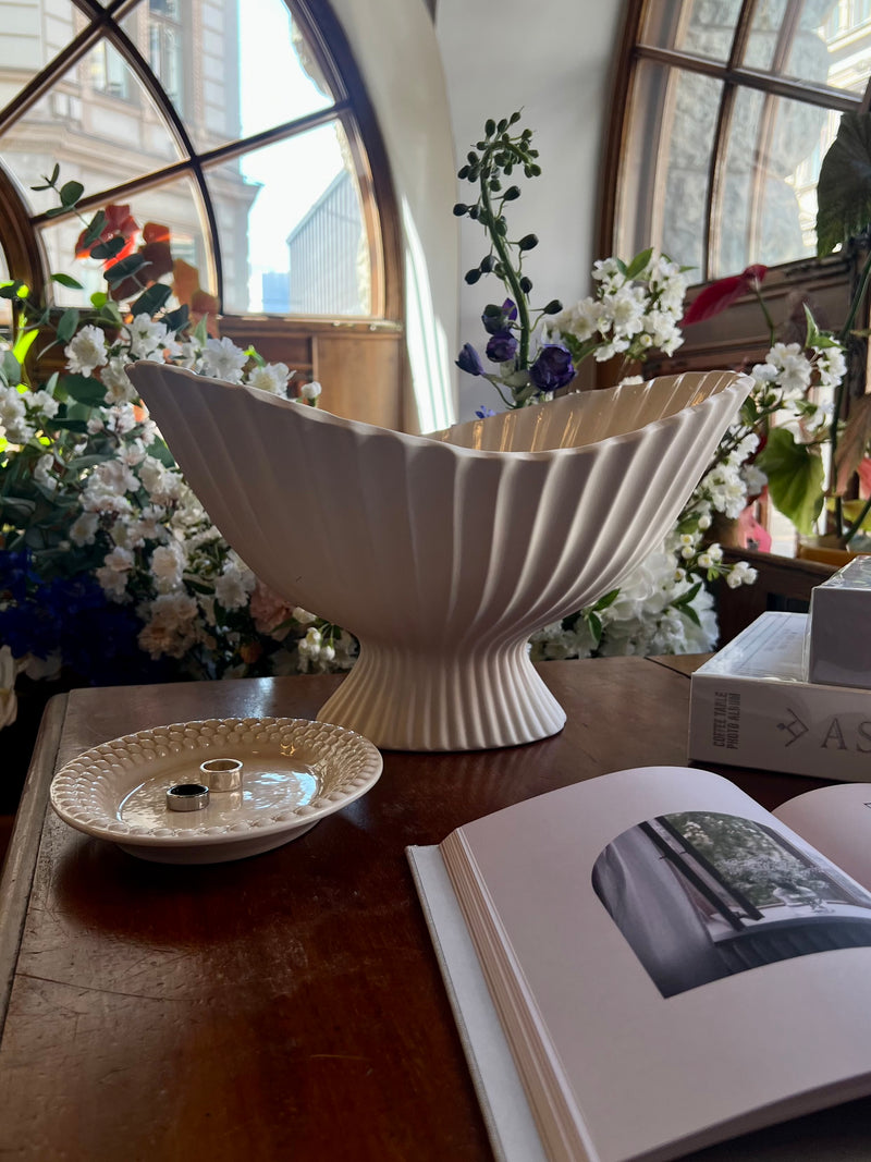 The Fountain from Ferm Living, a large decorative bowl paired with the Mateus oval Bubbles plate holding jewellery are placed on a table with flowers in the background and beautiful interior design book on the foreground.