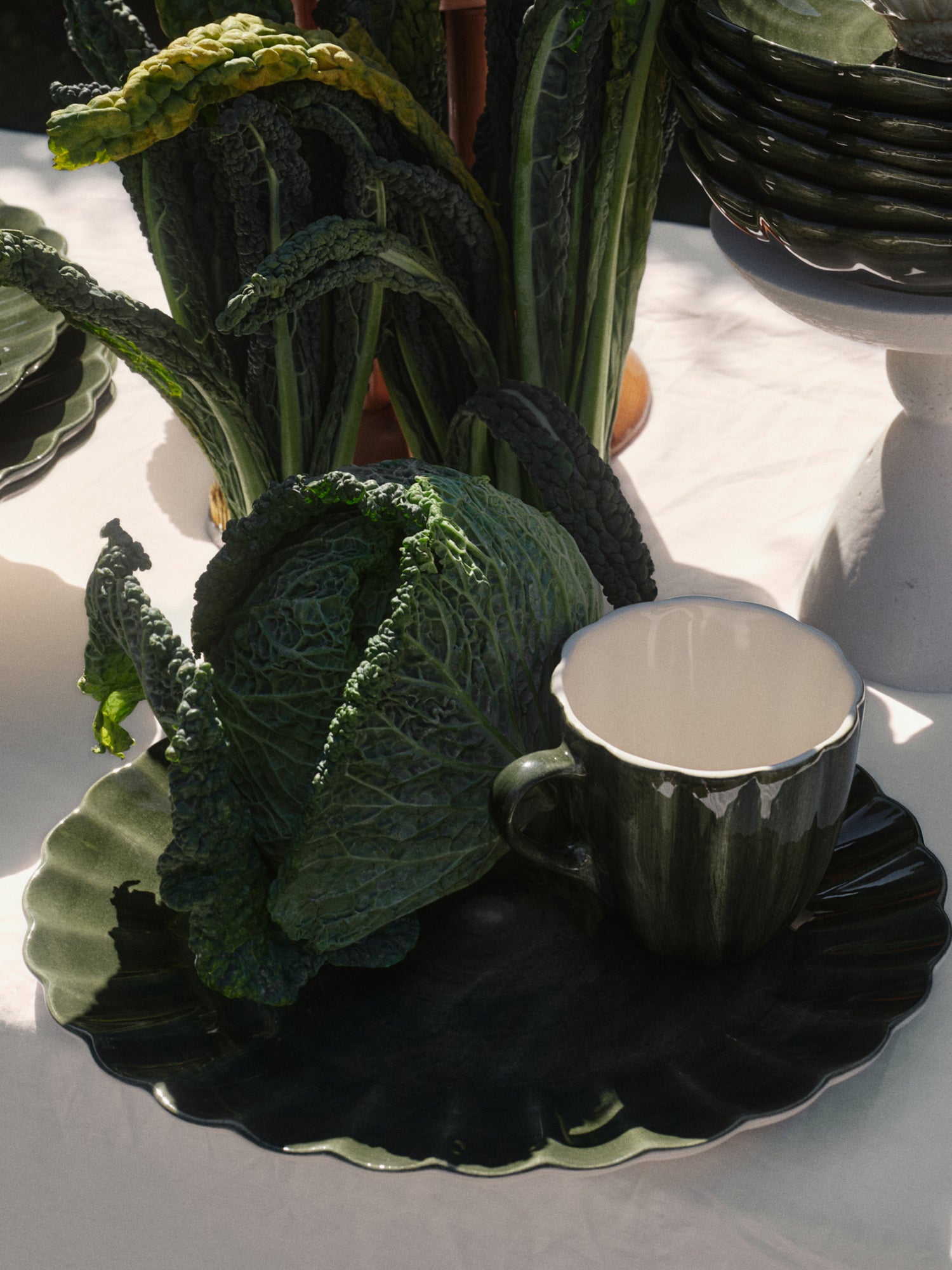 Decorative arrangement of green leafy vegetables and forest green Oyster collection oval platter, coffee mug and other plates on a white tablecloth.