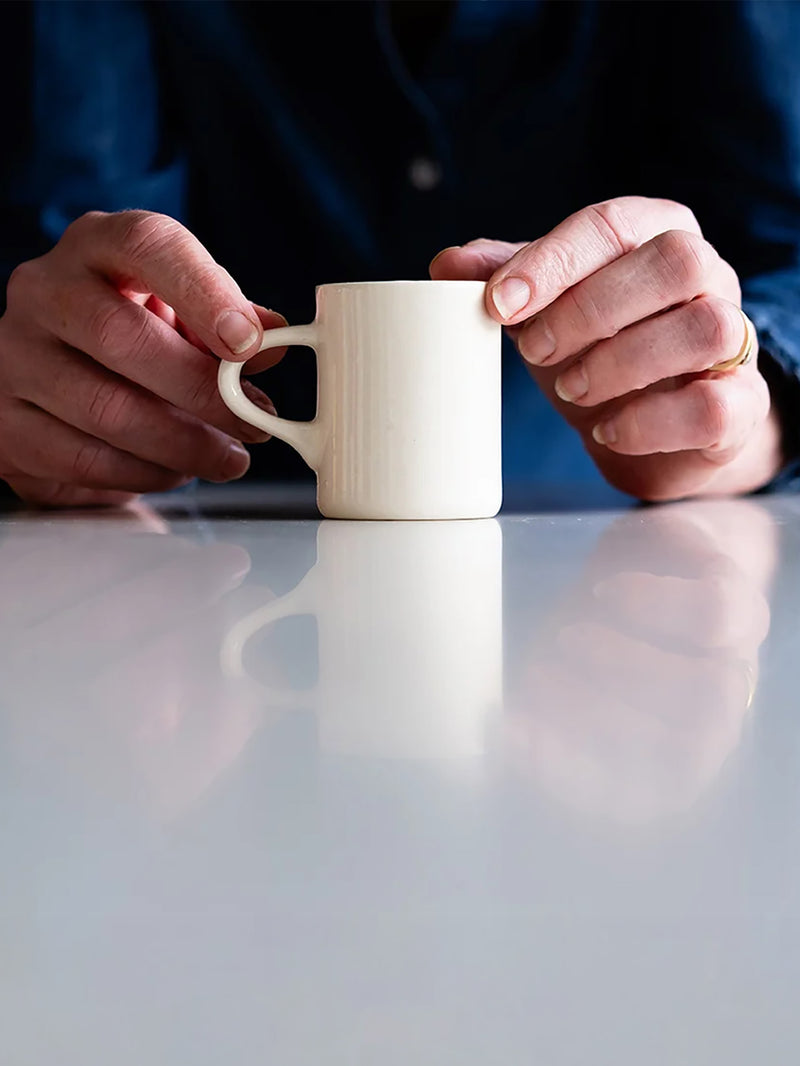 A small plain offwhite ceramic mug with a handle, held by two rugged hands over a reflective light gray surface. The person is wearing a dark blue denim shirt and a gold ring.