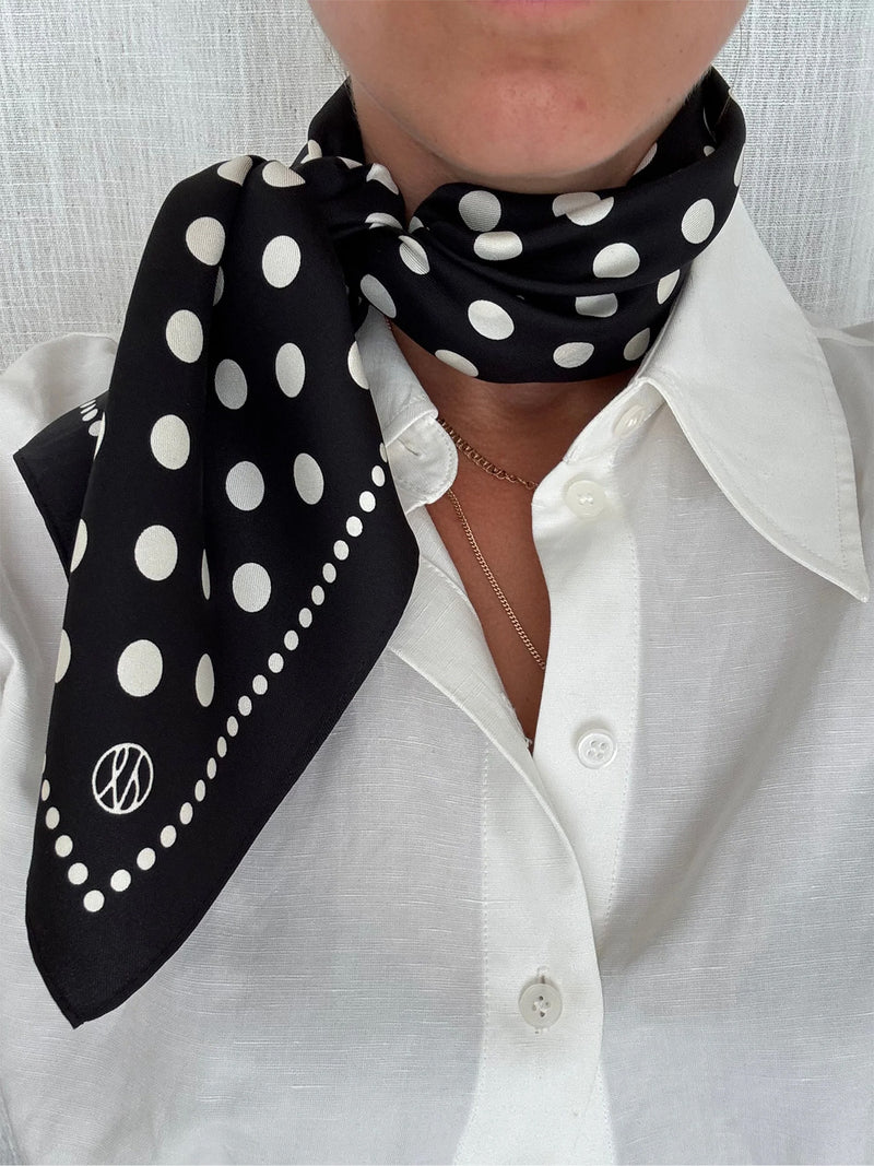 A close-up of a person wearing a black silk scarf with white polka dots and a small circular logo, tied around the neck over a white button-down shirt.
