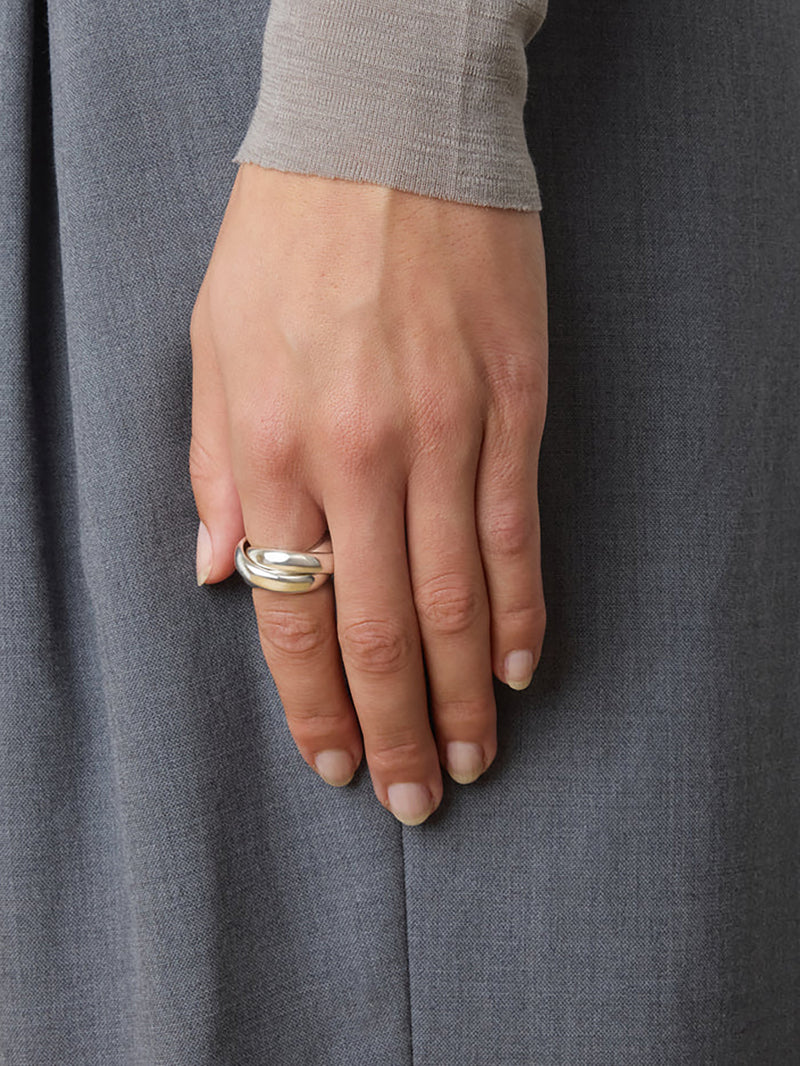 A close-up of a person's hand wearing a double-band ring in silver, set against a gray textured fabric trouser leg or skirt background.