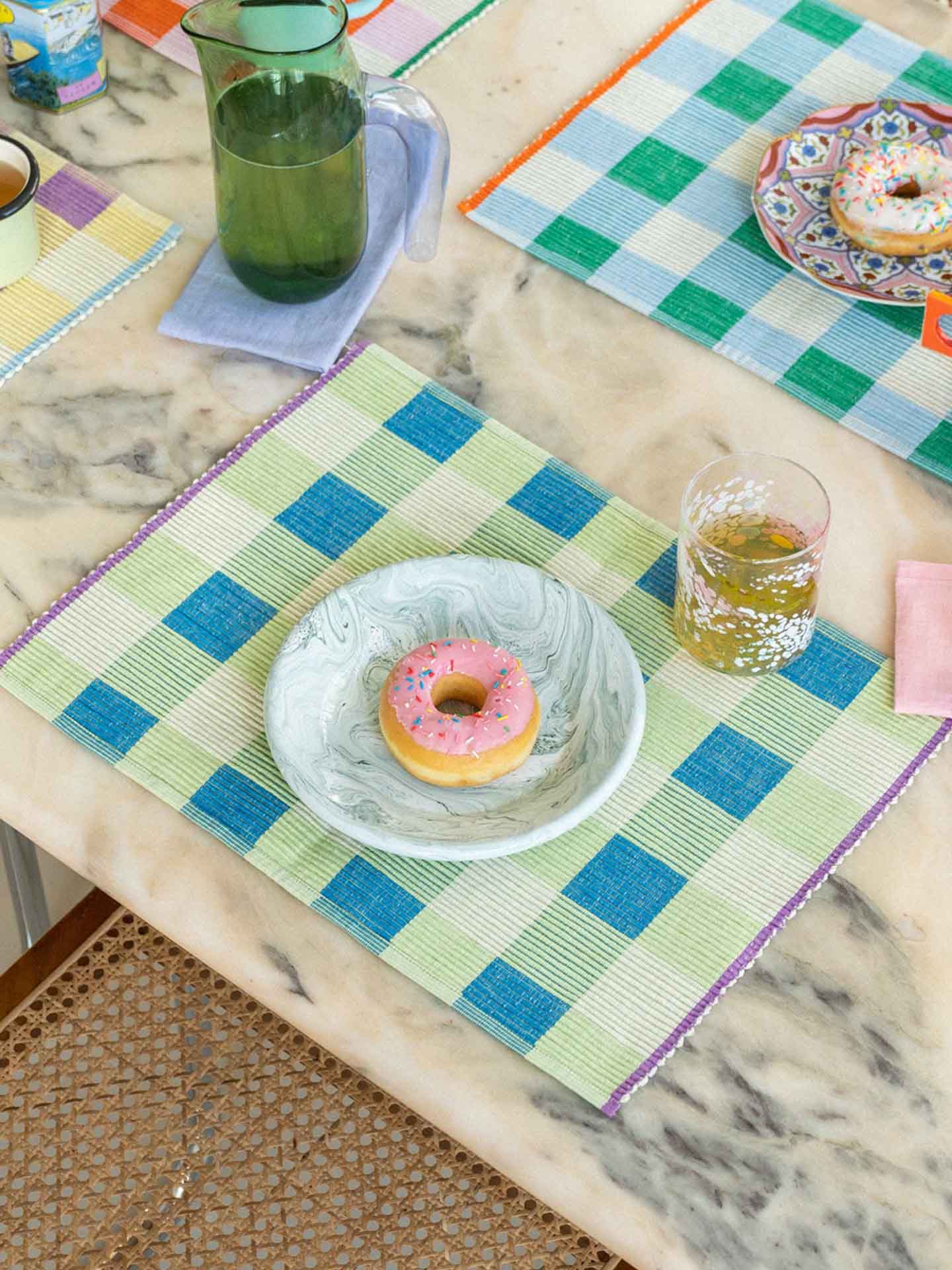 Table setting with colourful checkered with green, turquoise and purple placemats, donuts, and drinks on a marble dining table.