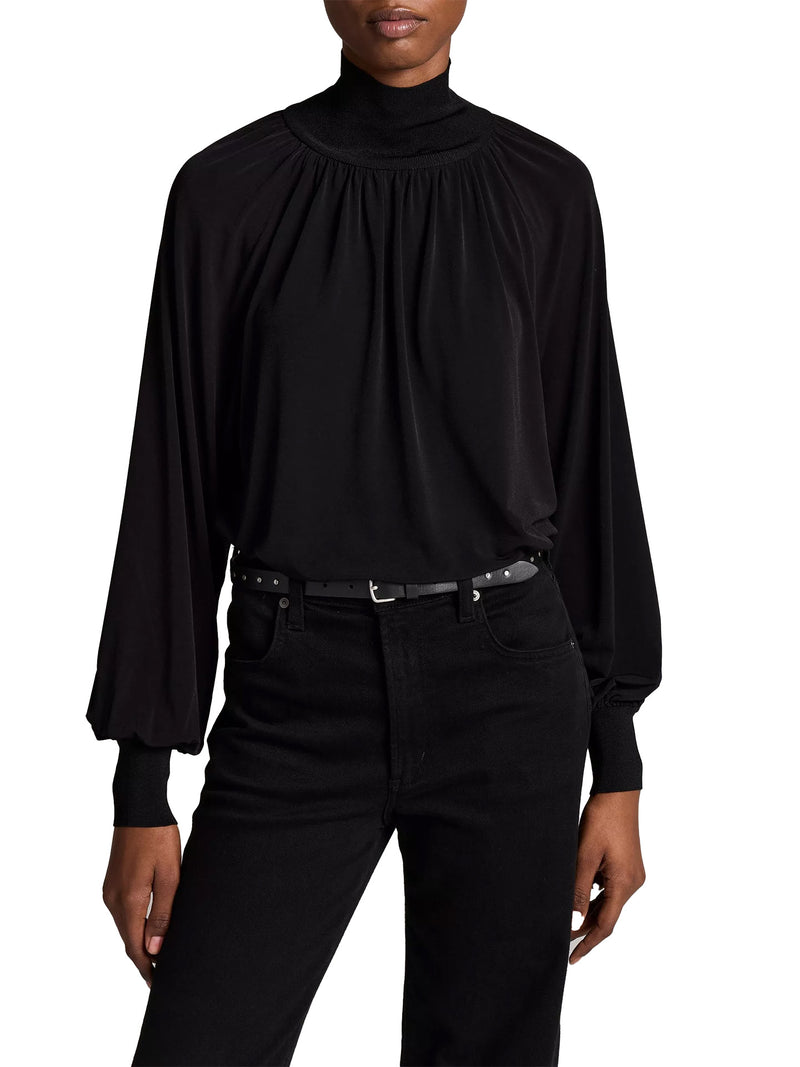 A model wearing a black long-sleeve mock neck blouse with a gathered, pleated yoke and black jeans, shown from the neck down against a plain white background.