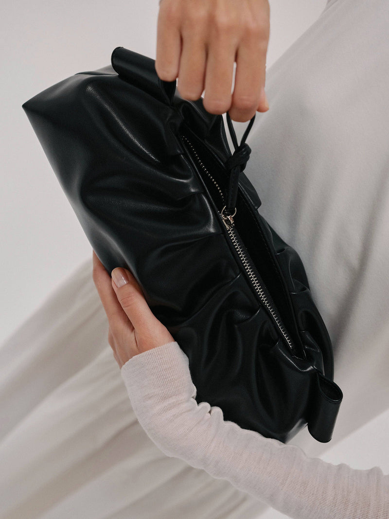 Black clutch bag held on one hand while closing the bag's zipper from a pull by a model wearing a white sweater against a neutral background