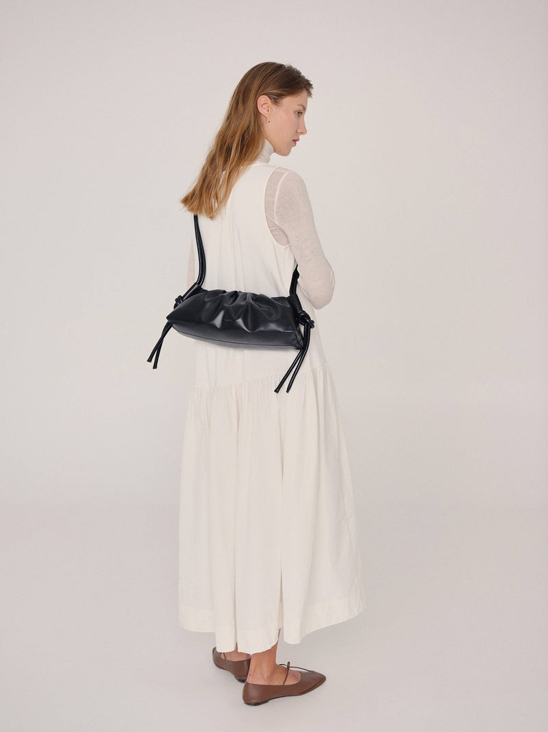 Woman wearing a small black handbag on a plain background across her shoulders as a crossbody bag.