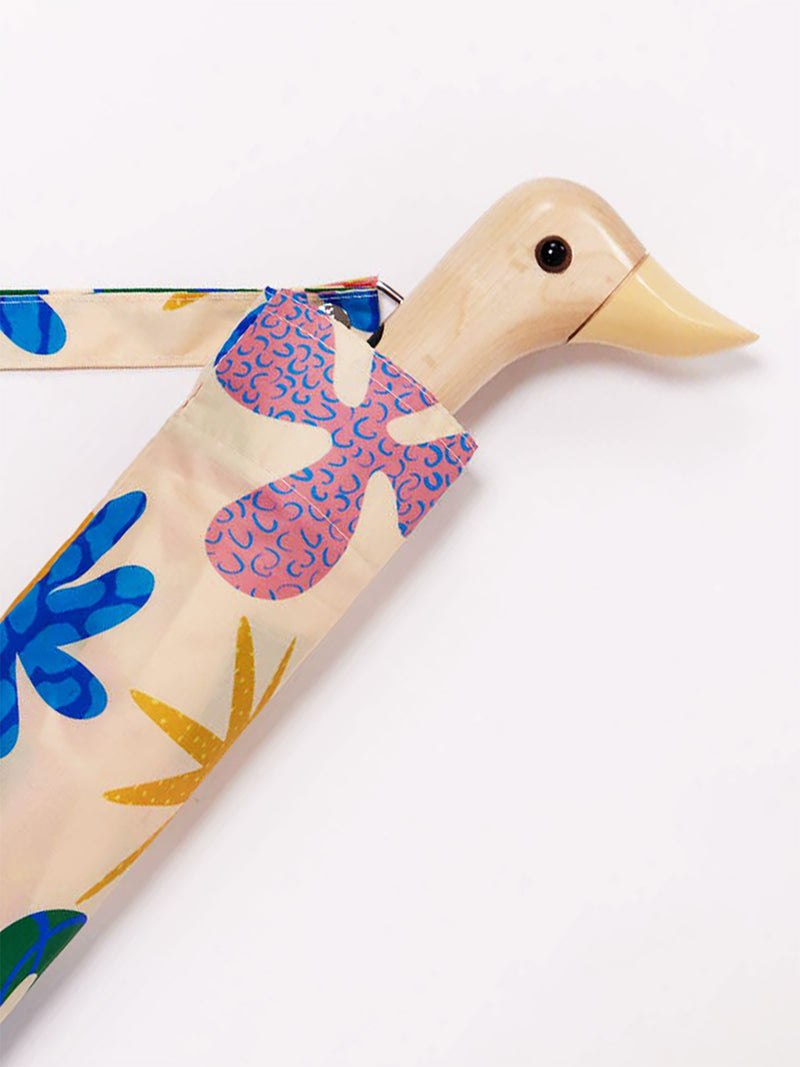 A wooden duck-shaped umbrella handle with a beige body, light butter yellow beak, and black eye, partially covered by a patterned fabric sleeve in blue, pink, and yellow on a cream background, set against a plain white studio background.