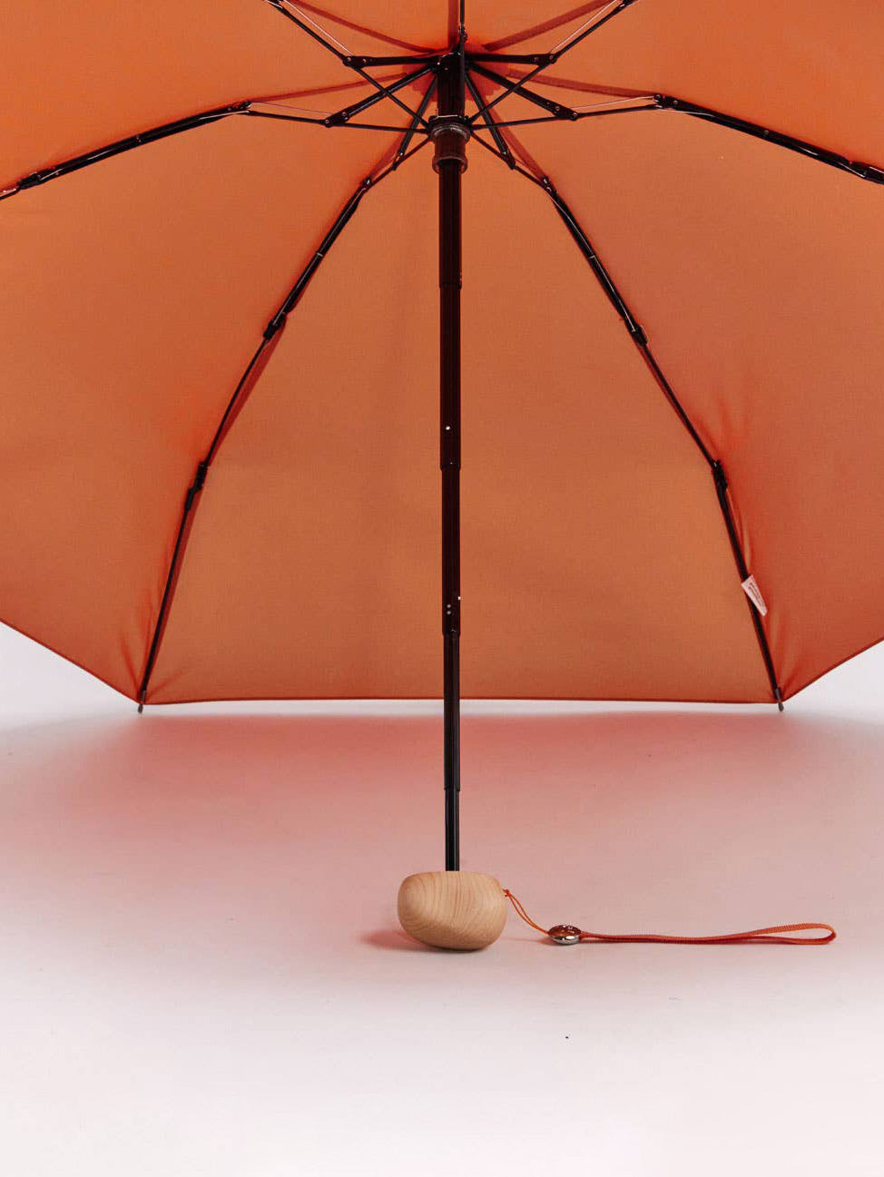 An dark orange umbrella shown open from below, with a black pole and ribs, a small light wood knob-shaped handle, and an orange wrist strap on a plain white studio background.