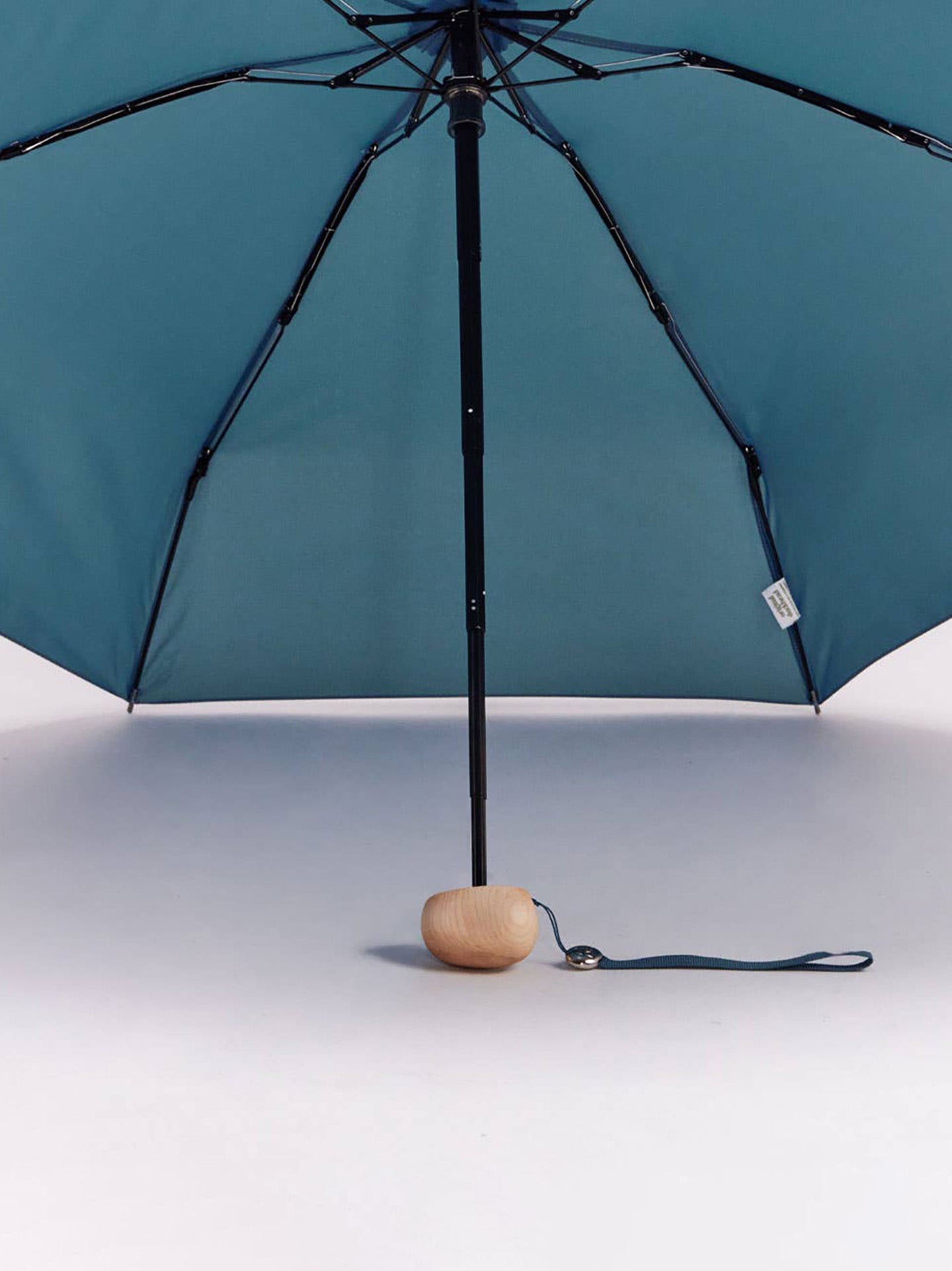 A teal blue folding umbrella with a black frame and a small oval wooden base, shown open against a clean white background.