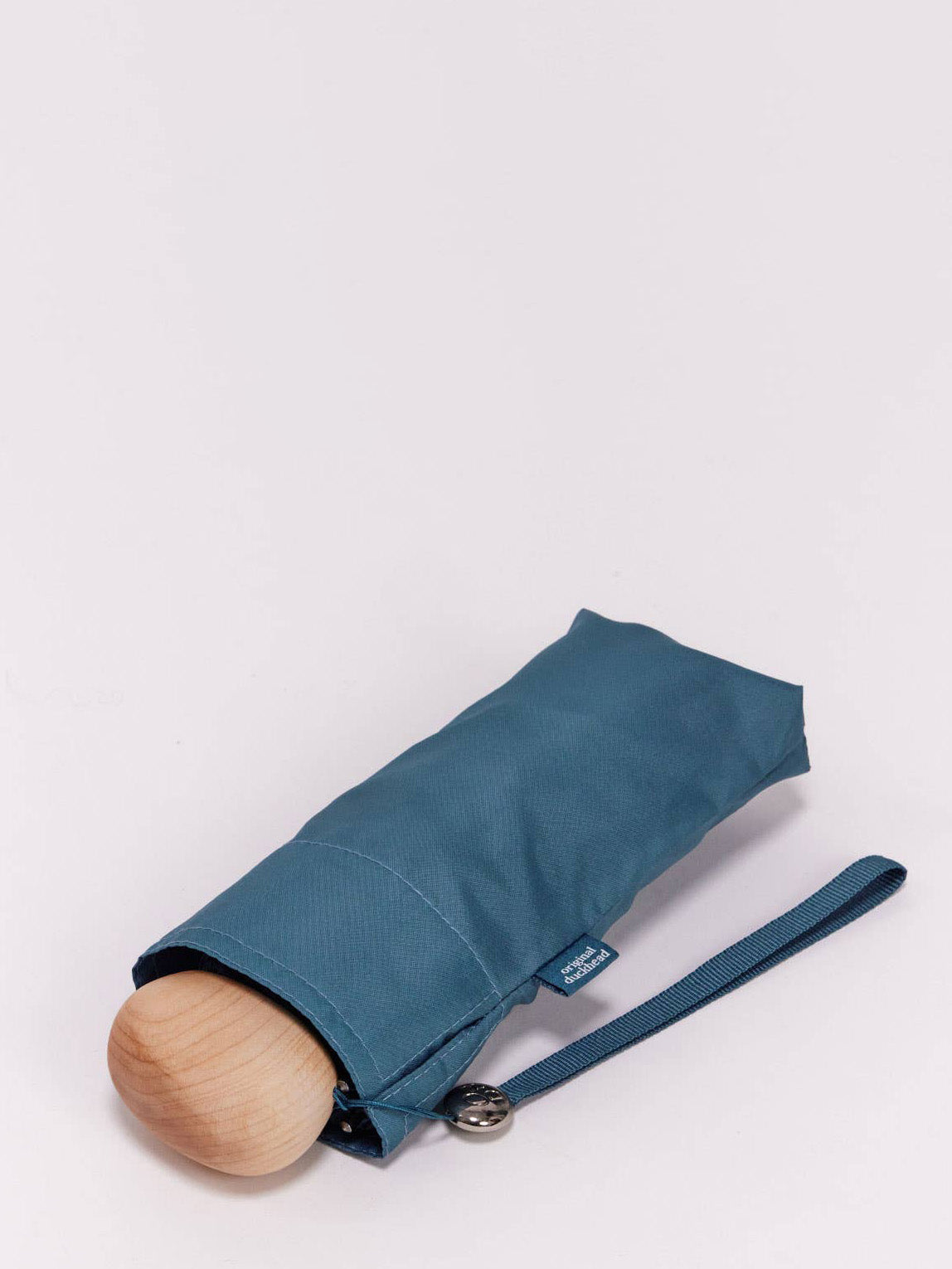 A very small folded teal blue umbrella closed in a matching fabric sleeve with a oval wooden handle and teal blue wrist strap, photographed on a clean white background.