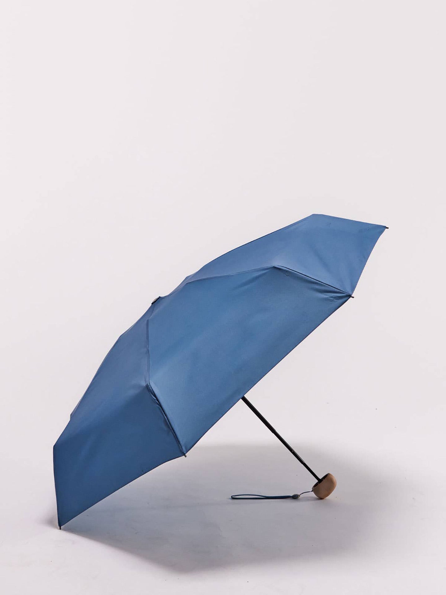 Open blue folding umbrella with a small light birch wood handle and wrist strap, photographed on a clean light gray studio background.