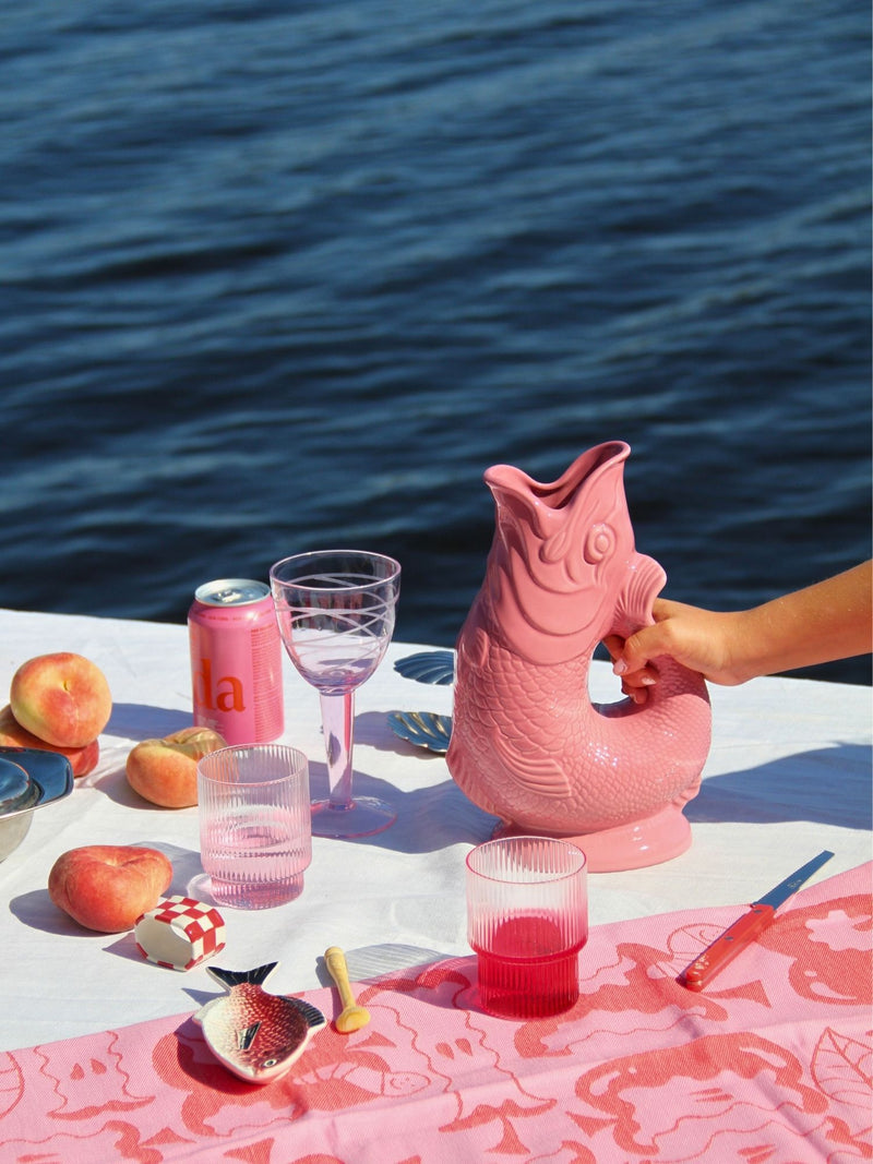 A pink fish-shaped pitcher sits on a white tablecloth outdoors by the water, surrounded by pink drinkware, peaches, and a hand reaching toward the pitcher.