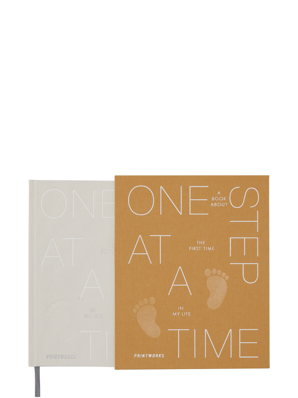 A baby book titled 'One Step at a Time' with a beige cover and a rusty yellow slipcase. The book's text is printed in a clear font, and it features images of a baby's feet, indicating the title's theme.