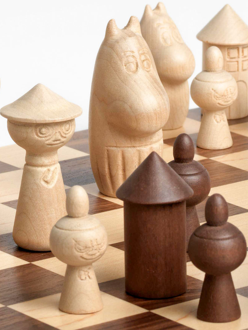 A set of uniquely shaped wooden chess pieces arranged on a wooden checkered chessboard. The pieces include stylized Snufkin, Snusmumriken in original story and Nuuskamuikkunen in Finnish translation, Moominmamma or Muumimamma in Finnish translation, Moomintroll, Muumipeikko in Finnish translation and and Little My, Lilla My in original story and Pikku Myy in Finnish, and the Moomin house figures in two natural wood tones: a light cream/beige tone and a darker brown tone.
