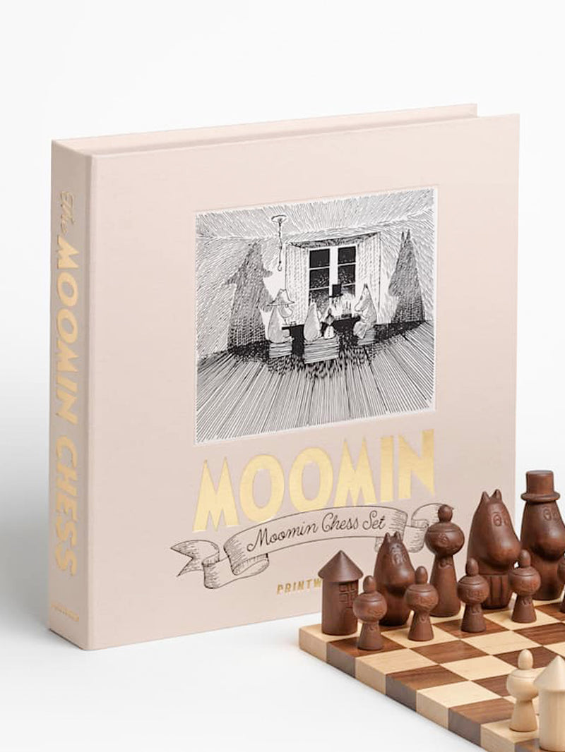 Moomin Chess set in a pale blush pink hardcover box with gold lettering, shown next to a wooden chessboard and chess pieces arrangent in four rows, Moominpappa playing as the king, Moominmamma as the queen, Mymlan as the bishop, Moomintroll as the knight and Moominhouse placed on the corner as the tower, with Little My figurines on the front row serving as pawns, on a white background.