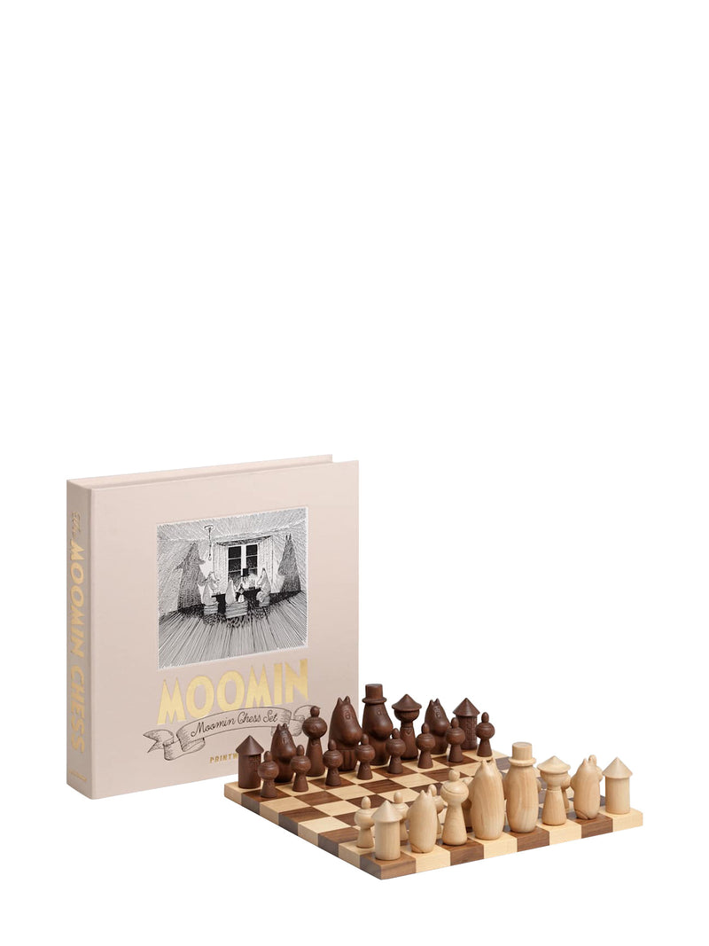 A wooden chess set with two-tone light and dark brown pieces arranged on a checkered board ready for play, next to a beige box titled "MOOMIN" with gold and smaller title "Moomin Chess Set" on an illustrated banner, with a large illustration from the Moomin books or comics by Tove and Lars Jansson in black-and-white on the cover, photographed on a white background.
