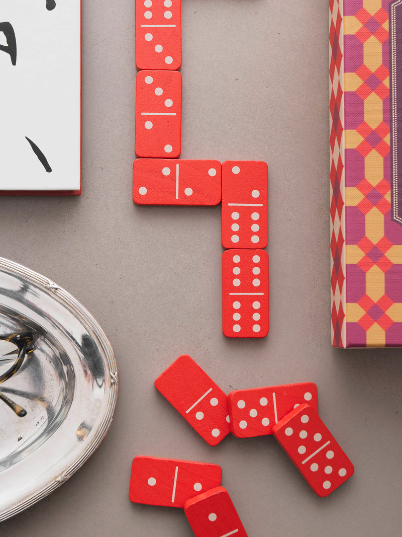 Red domino tiles with white pips arranged on a neutral gray surface, alongside partial views of patterned and white boxes and a silver dish.