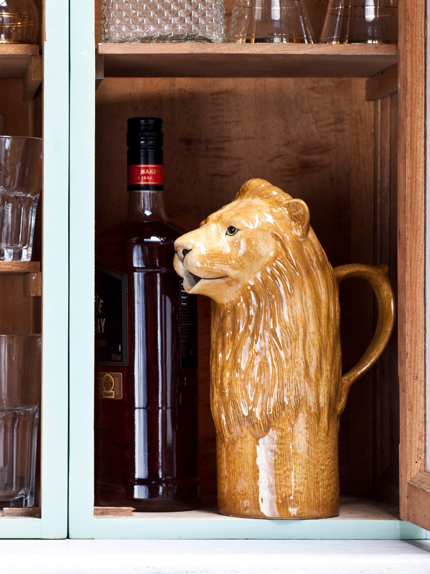 Ceramic water jug shaped like a lion's head on a rustic cupboard shelf with a wine bottle and glasses in the background